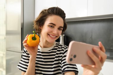 Gülümseyen, güzel genç bir kadın taze sarı biberle selfie çekiyor, yemek blogcusu mutfakta sebzelerle selfie çekiyor, vegan sosyal medya için yemek yapıyor ve video kaydediyor..