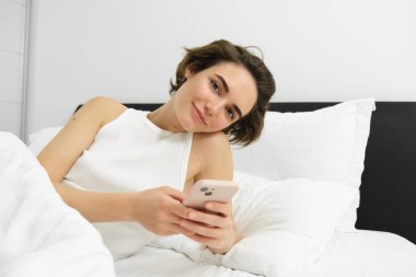 Gülümseyen güzel bir kadın, akıllı telefonuyla yatakta uzanıyor, cep telefonuna bakıyor, uygulamayı kullanıyor, mesajları okuyor ve memnun görünüyor..