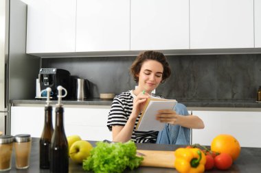 Mutfakta gülümseyen genç bir kadının portresi, elinde defter, yemek tarifi için notlar almak, market listesi yazmak, salata pişirmek, sebzelerin yanında oturmak ve kesim tahtası..
