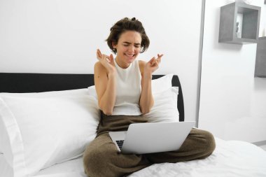 Yatak odasında bir kadının portresi, yatağında oturuyor, dizüstü bilgisayar kullanıyor, iyi haber bekliyor, dua ediyor, dilek tutuyor, bir şeyler bekliyor, olumlu sonuç için dua ediyor..