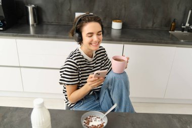 Çay içen ve kulaklıkla müzik dinleyen genç bir kadının portresi, mutfakta yemek yerken sosyal medyayı kaydırmak, mutlu bir şekilde gülümsemek..