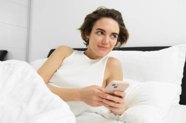 Güzel, esmer, genç bir kadının uyandığı, yatakta yattığı, akıllı telefonu tuttuğu ve gülümsediği, uyku oranını kontrol ettiği görüntüsü..