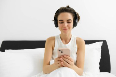Yatakta yatan, kablosuz kulaklık takan, akıllı telefona bakan, internetten video izleyen, yatak odasında cep telefonu kullanan genç bir kadının portresi..