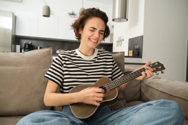 Genç modern kadın portresi, evde gitar çalan öğrenci, küçük bir gitarla oturan, şarkı söyleyen ve mutlu hisseden, kanepede oturan. Yaşam tarzı ve müzik konsepti