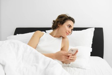 Yatakta beyaz çarşaflara uzanmış, akıllı telefon kullanan, cep telefonu tutan bir kadının portresi. Yaşam tarzı ve teknoloji kavramı