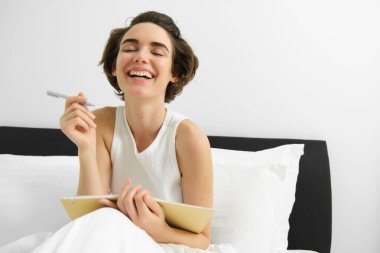 Not defteri ve kalemle güzel bir kadının portresi, yatakta oturuyor, gülüyor ve gülümsüyor, günlüğe yazıyor, yatak odasında çalışıyor..