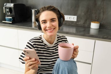 Çay içen ve kulaklıkla müzik dinleyen genç bir kadının portresi, mutfakta yemek yerken sosyal medyayı kaydırmak, mutlu bir şekilde gülümsemek..