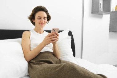 Tarz sahibi, modern bir kadının portresi, cep telefonu tutan, gülümseyen ve akıllı telefona bakan, yatakta uzanan, evinde dinlenen.