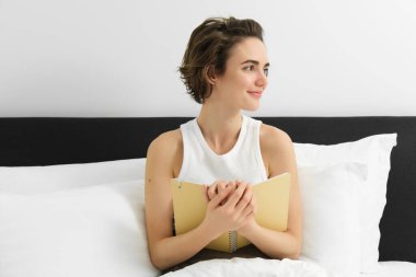 Genç şehvetli bir kadının portresi, defterine sarılıyor, günlük ya da günlük tutuyor, gülümsüyor ve yan bakıyor, yatakta oturuyor, rüyalarını ya da duygularını not ediyor..