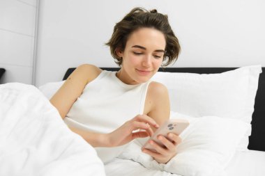 Yatakta kadın modelin resmi, akıllı telefona bakıyor, mesaj gönderiyor, cep telefonunda çalar saati açıyor, yatak odasında uyuyor..