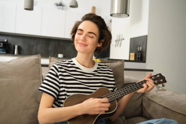 Güzel gülümseyen bir kadının yakın portresi, ukulele çalmak, kapalı gözlerle telleri tıngırdatmak ve mutlu bir yüz, oturma odasında oturmak..