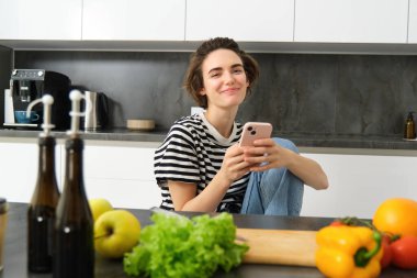 Akıllı telefonlu esmer bir kadının portresi mutfakta oturmuş, yemek tarifi videoları izliyor, doğrama tahtası ve sebzeleri var..
