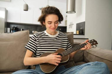 Genç modern kadın portresi, evde gitar çalan öğrenci, küçük bir gitarla oturan, şarkı söyleyen ve mutlu hisseden, kanepede oturan. Yaşam tarzı ve müzik konsepti