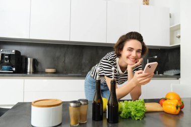 Kadın portresi, yaşam tarzı blogcusu, akıllı telefon uygulamasında yemek pişirme sürecinin resmi, sebzelerin yanında durmak ve kesim tahtası..