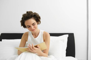 Günlüğüyle yatakta oturan bir kadının portresi, sabahları düşünceleri ve duyguları, elinde defter tutarak gülümsüyor..