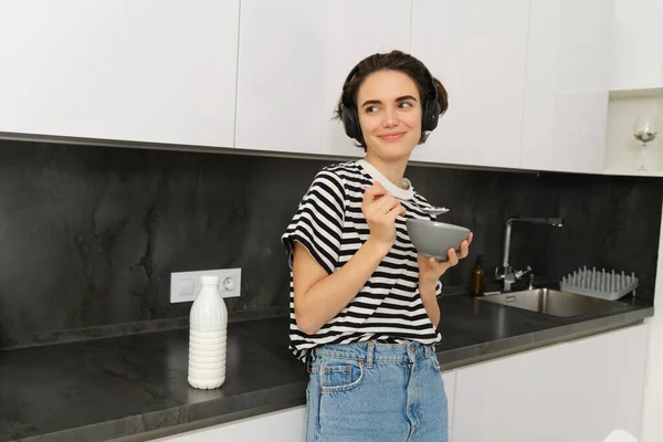 Portrait of modern woman eats breakfast, holds bowl and spoon, stands in kitchen, listens music in wireless headphones. Concept of lifestyle and home.