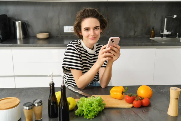 Modern genç bir kadının portresi ne pişireceğini düşünüyor, akıllı telefon ve sebzelerle mutfakta oturuyor, yemek yapıyor, sağlıklı diyet salatası yapıyor..