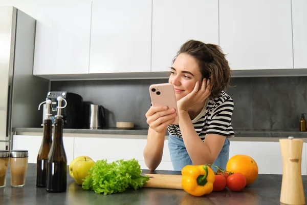 Portrait of young smiling woman, searching cooking recipe online on smartphone, standing near vegetables and chopping board, making meal, preparing salad and using social media on mobile phone.