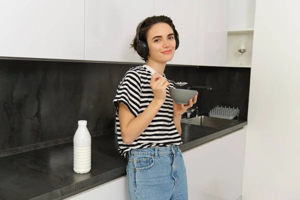 Portrait of modern woman eats breakfast, holds bowl and spoon, stands in kitchen, listens music in wireless headphones. Concept of lifestyle and home.