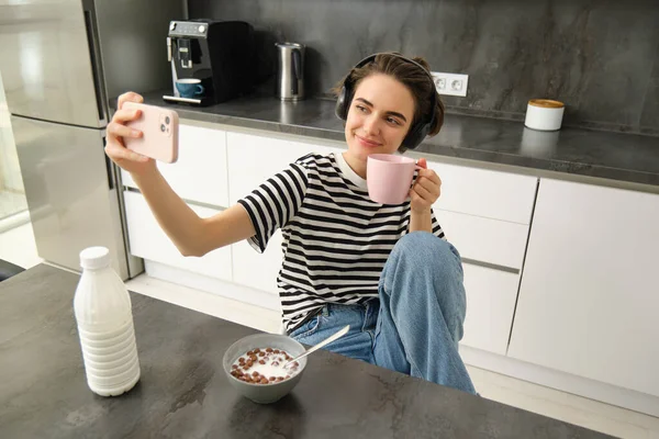 Portrait of modern happy woman, taking selfie, lifestyle blogger recording video of her eating breakfast and drinking tea in the kitchen, using smartphone for making content.