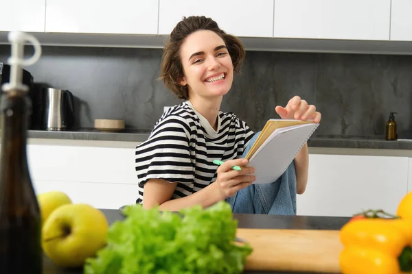 Portrait of young woman cooking, writing notes, grocery list in notebook, creating list of meals to cook through meal, sitting in kitchen near vegetables and chopping board.