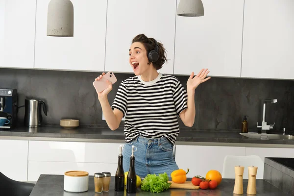 Portrait of carefree woman singing in smartphone mic, listening music in headphones while cooking breakfast, making a meal, chopping vegetables and dancing.