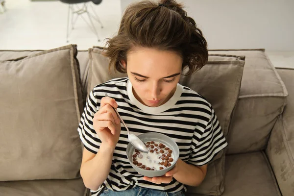 Upper angle shot of happy, cute young woman on sofa, eating bowl of cereals with milk and smiling, enjoying her breakfast.