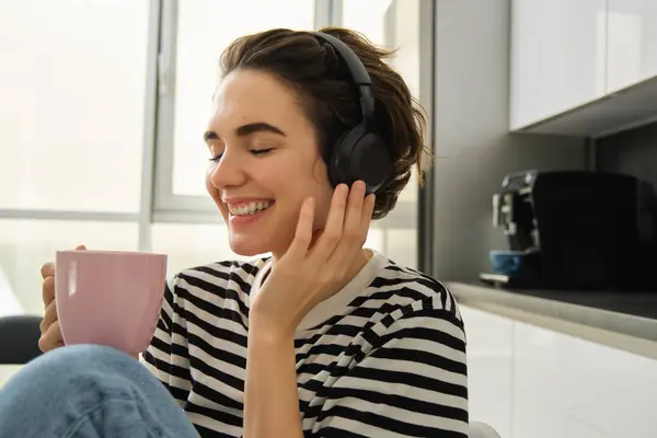Lifestyle and music concept. Happy smiling woman with cup of tea, sits in kitchen and touches headphones on her head, listens to music with pleased relaxed face.