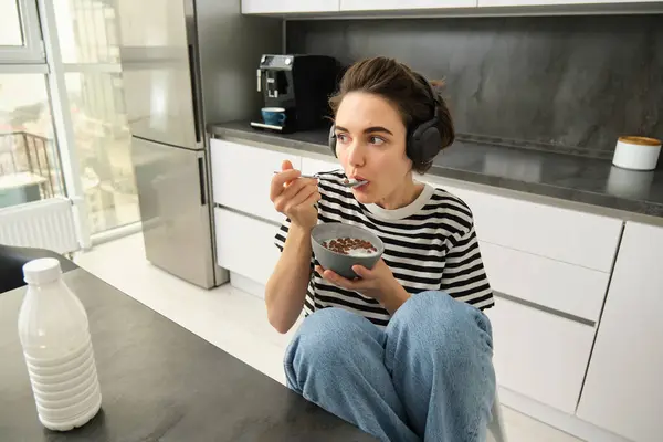 Image of stylish, modern woman, student eating cereals with milk for breakfast, holding bowl and spoon, listening music in wireless black headphones.