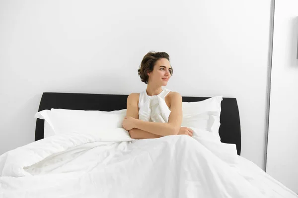 Portrait of young smiling woman with messy hair, sitting on her bed, covered in white linen sheets, looking aside with dreamy face expression, waking up in morning.