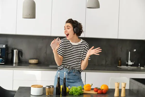 Portrait of woman singing and dancing while cooking food in the kitchen, holding smartphone, chopping vegetables on kitchen counter.