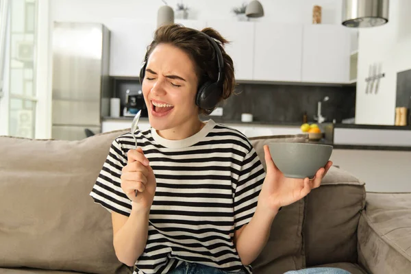 Young brunette woman holding bowl of cereals, singing with spoon in hand, listening to music in headphones, eating breakfast and enjoying favourite song, sitting on sofa at home.
