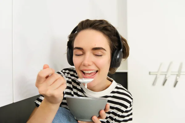 Close up portrait of young woman enjoys eating her breakfast, having cereals with milk, holding spoon and bowl, sits on kitchen counter with headphones, listens to music.