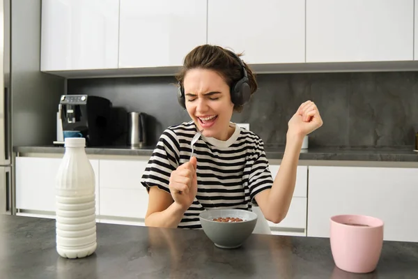 Happy and emotional young woman, singing while eating breakfast, having her cereals with milk, listening to music in wireless headphones, sitting in the kitchen.