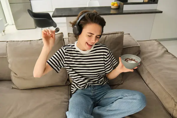 Portrait of stylish young woman listening to music in headphones, eating cereals with milk on sofa in living room, dancing while sitting and having breakfast, holding bowl and spoon.