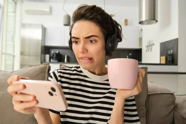 Portrait of woman with scared, concerned face watching video on smartphone, drinking tea, wearing headphones, sitting on sofa at home.