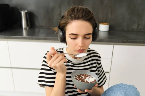 Image of stylish, modern woman, student eating cereals with milk for breakfast, holding bowl and spoon, listening music in wireless black headphones.