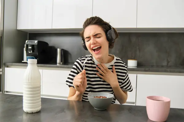Carefree young woman, student in headphones, eating cereals for breakfast, listening music, singing and using soon as a microphone, having fun while having her meal in the morning.