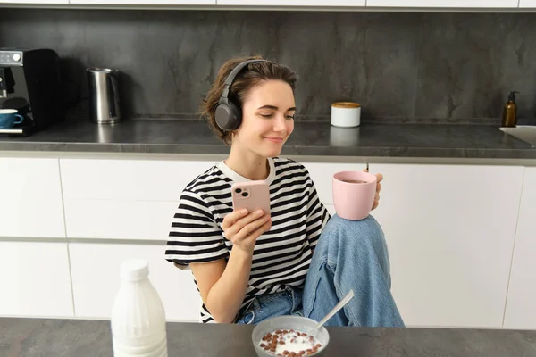 Stylish young woman in headphones, listens music and uses smartphone, drinks warm tea in kitchen, relaxing at home.