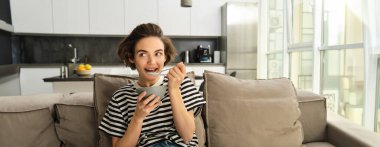 Kahvaltı eden, kanepede sütlü gevrek yiyen, gülümseyen ve tatmin olmuş görünen genç bir kadının portresi..