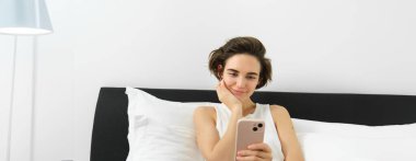 Yaşam tarzı ve teknoloji konsepti. Gülümseyen güzel bir kadın akıllı telefonuyla yatakta uzanıyor, yatak odasında dinleniyor, sosyal medyada sohbet ediyor, online alışveriş yapıyor..