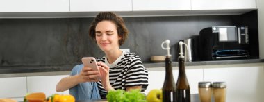 İnternette yemek tarifi arayan genç bir kadının portresi akıllı telefon kullanıyor, sebzelerin yanında oturuyor, salata malzemelerinin yanında oturuyor ve kameraya gülümsüyor..