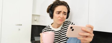 Akıllı telefon ekranında üzgün suratlı, sempatik bir kadın portresi, cep telefonu uygulamasında video izleme, mutfakta çay ya da kahve içme, kaşlarını çatma ve acımayla bakma.