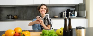 Gülümseyen, gülen ve akıllı telefon kullanan genç bir kadının portresi yemek pişirirken, akıllı telefon uygulamasında yemek tarifleri ararken, evdeki mutfağında sebzelerden yemek hazırlarken..