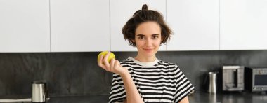 Genç kadın modelin portresi, mutfakta poz veriyor, bir elma tutuyor, gülümsüyor ve kameraya bakıyor..