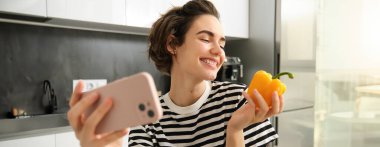 Gülümseyen modern kadın, vejetaryen yemek blogcusu, taze sarı biberli selfie çeken, akıllı telefon tutan, sosyal medya hesabı için bir video blogu kaydeden, mutfakta poz veren.