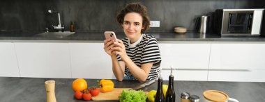 Modern genç bir kadının portresi ne pişireceğini düşünüyor, akıllı telefon ve sebzelerle mutfakta oturuyor, yemek yapıyor, sağlıklı diyet salatası yapıyor..