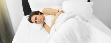 Karın ağrısı ile yatakta yatan kadının portresi, adet sancıları, kaşları çatık ve ağrı periyodu var. Refah ve sağlık kavramı