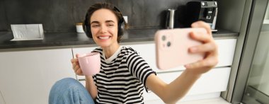 Modern mutlu kadın portresi, selfie çekiyor, yaşam tarzı blogcusu kahvaltısını kaydediyor ve mutfakta çay içiyor, akıllı telefonu içerik yapmak için kullanıyor..