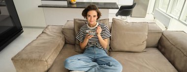 Gülümseyen kadın modelin üst açı çekimi, oturma odasında kanepede bir kase sütlü mısır gevreğiyle oturmak, televizyonun önünde yemek yemek, kahvaltısını yapmak ve dinlenmek..